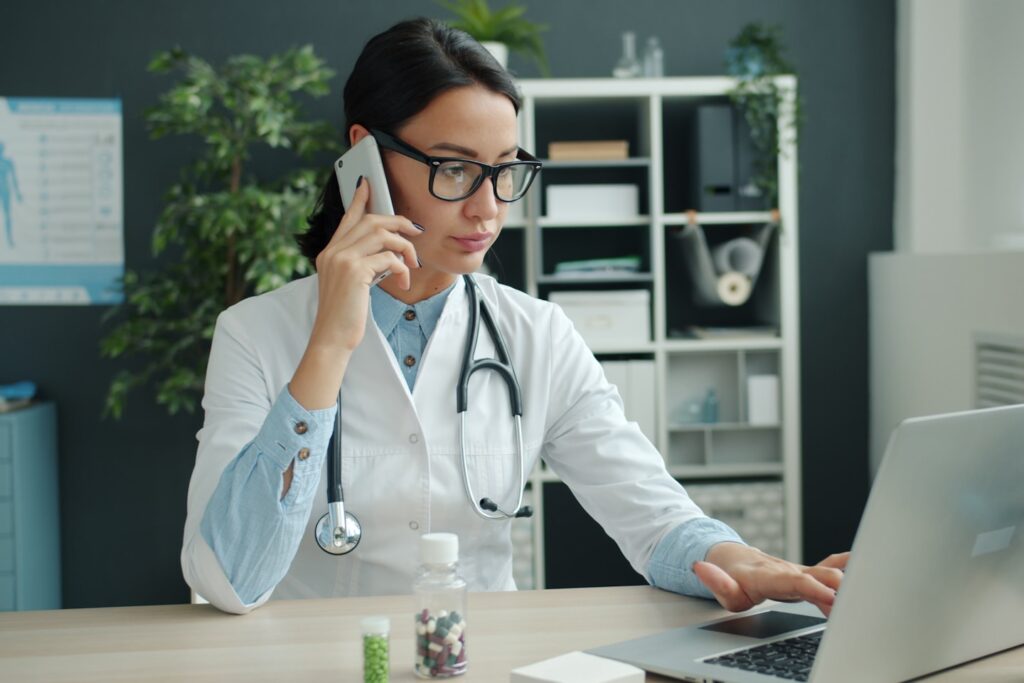 Young female doctor is talking on mobile phone and using laptop computer working in office alone. Modern devices, medicine and communication concept.